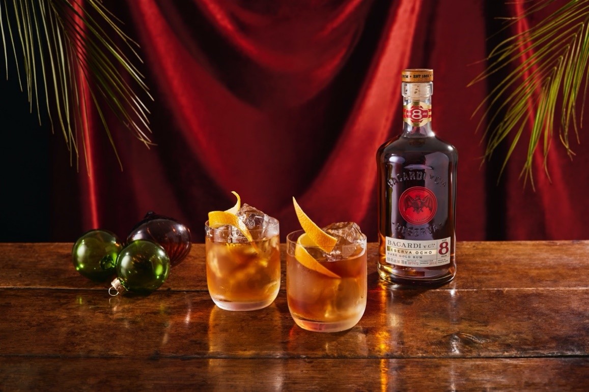 Bacardí Reserva Ocho rum bottle with two cocktails, orange twists, Christmas baubles.