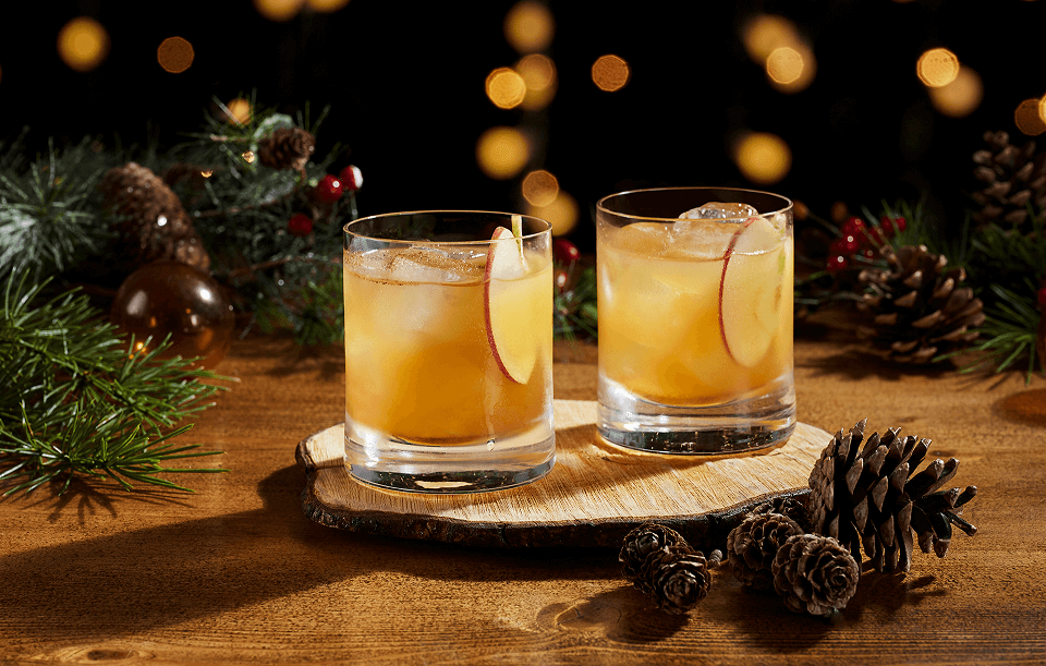 Two amber cocktails in rocks glasses, garnished with apple slices near pine cones and berries.
