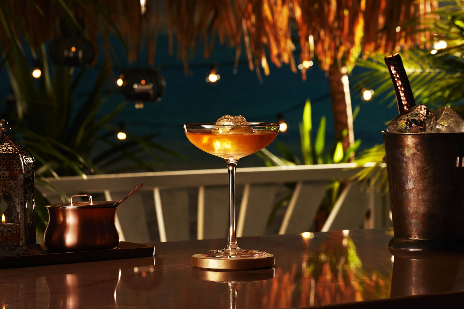 Amber cocktail in a coupe glass with a large ice cube at a tropical nighttime bar.
