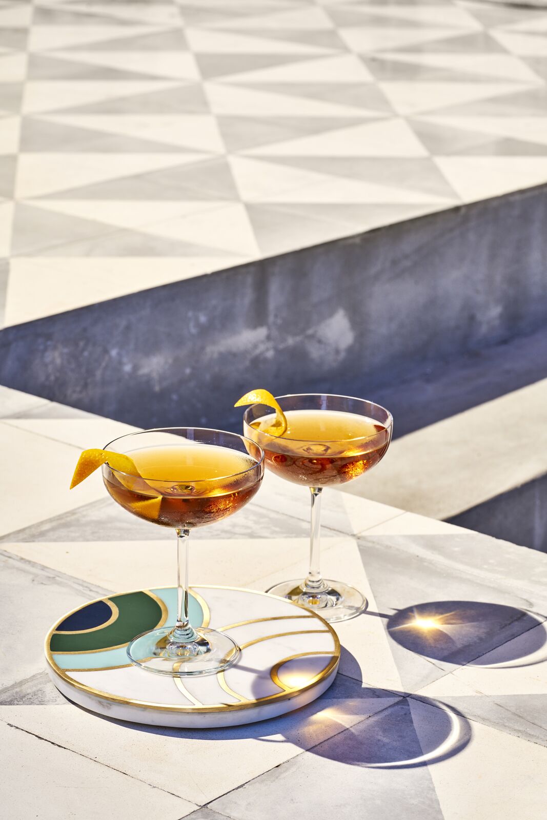 Two amber cocktails in coupe glasses, garnished with orange twists on decorative coasters.