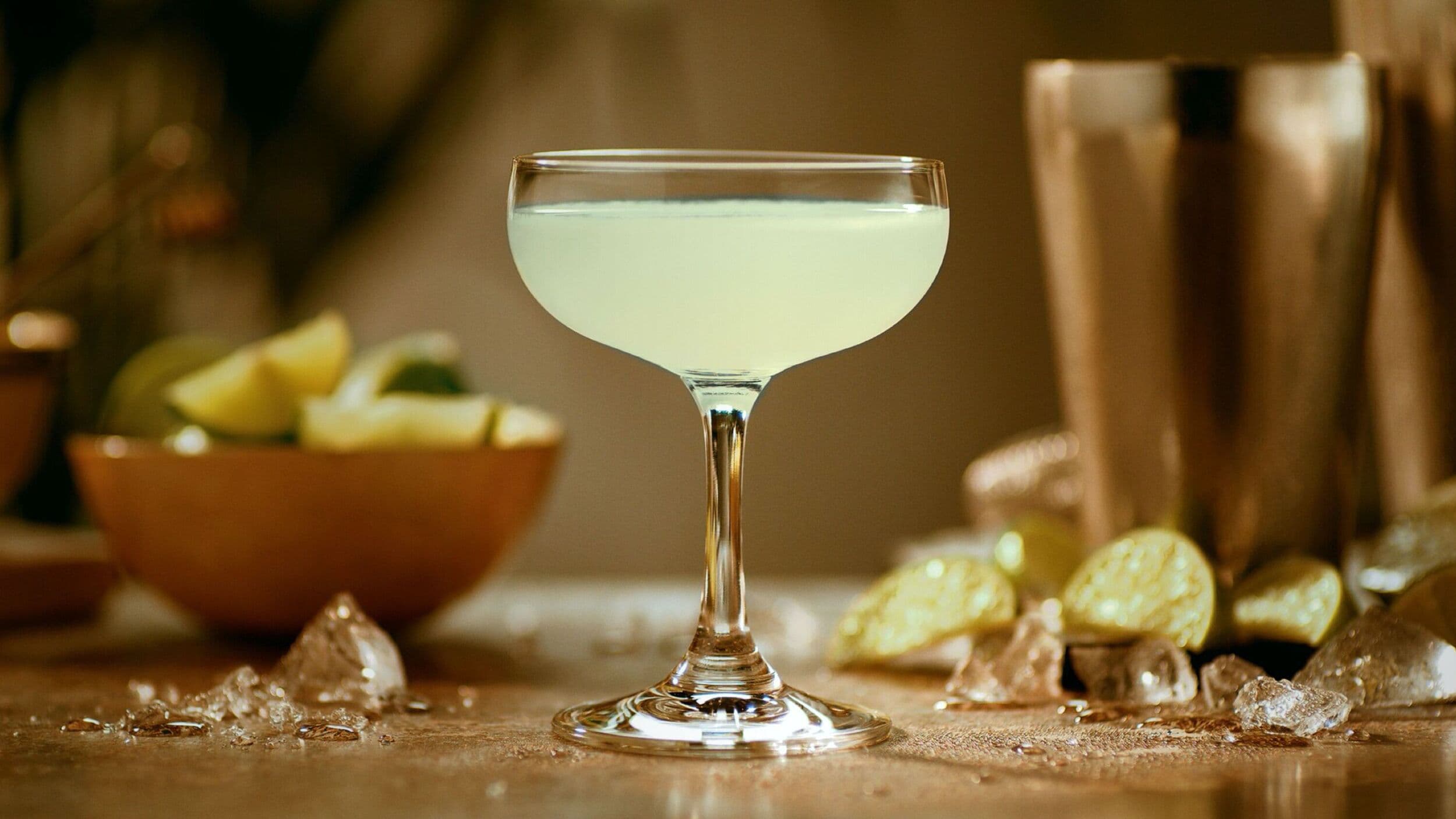 Pale yellow-green daiquiri cocktail in a coupe glass with scattered lime wedges and ice chunks.