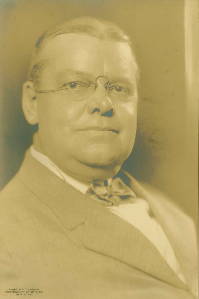 Sepia-toned vintage portrait of a man in a light suit jacket, white shirt, bow tie, wire-rimmed glasses.