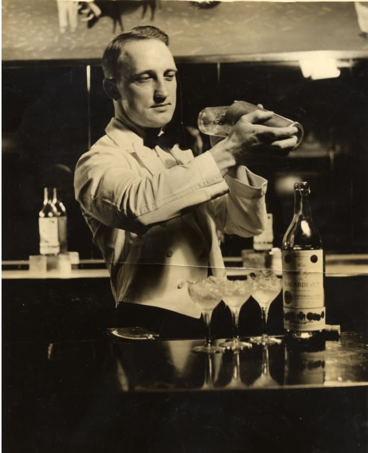 Black and white photograph of a bartender shaking a cocktail with BACARDÍ rum bottle nearby.