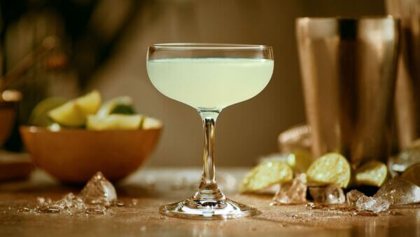 Light yellow-green cocktail in coupe glass, surrounded by lime wedges, ice cubes, cocktail shaker.