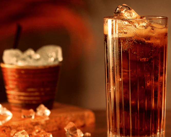 Tall glass of dark soda with ice, foam, and scattered ice cubes.
