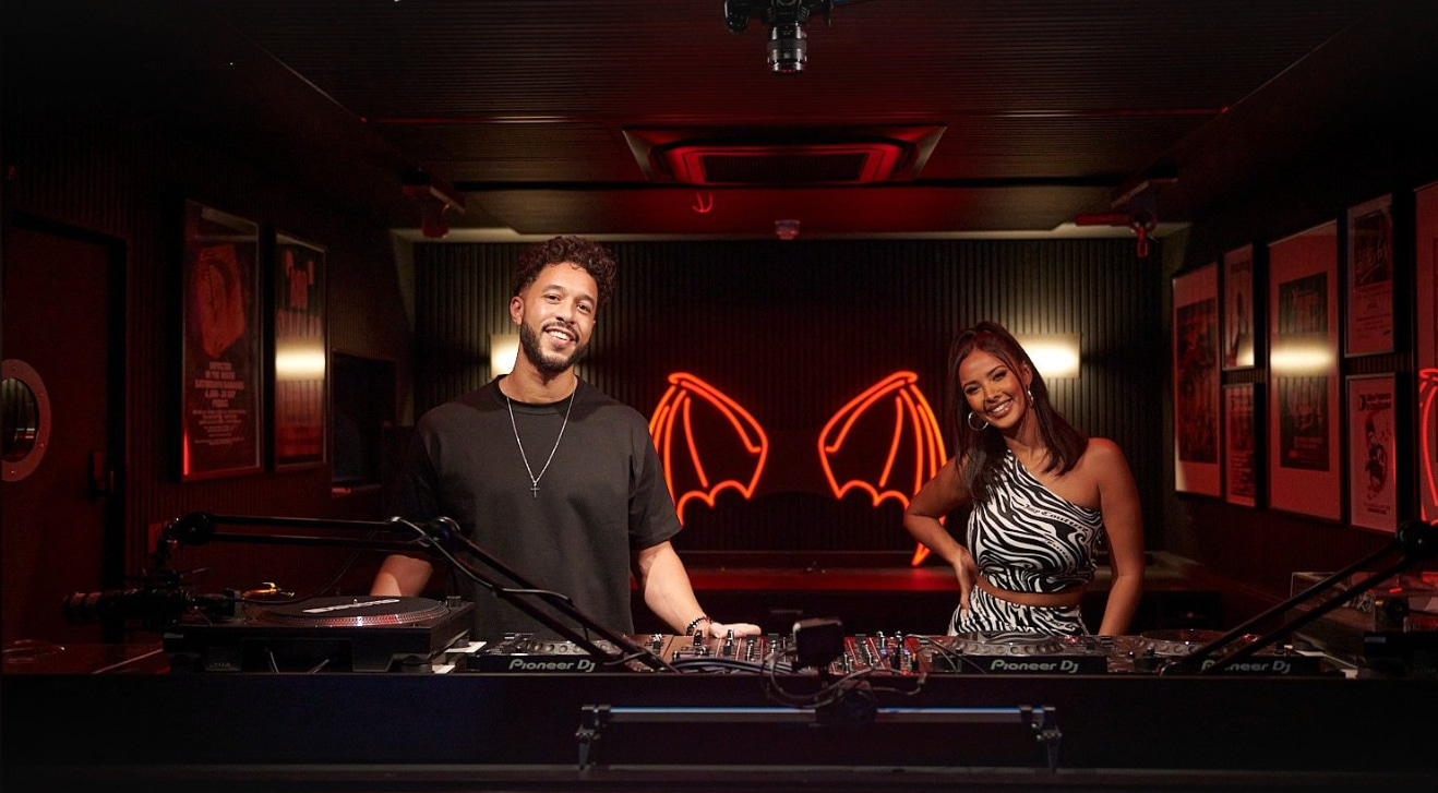 Two people standing behind a professional DJ setup with a glowing red neon bat wing sign.