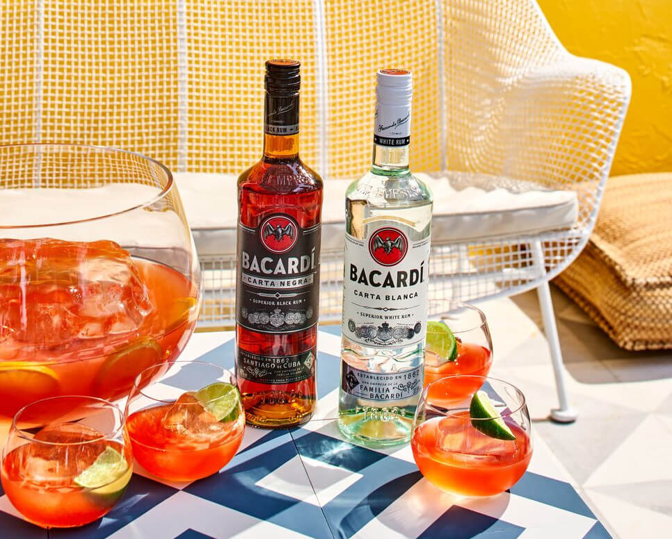 Two BACARDÍ rum bottles near punch bowl, cocktail glasses with lime garnishes, ice cubes.