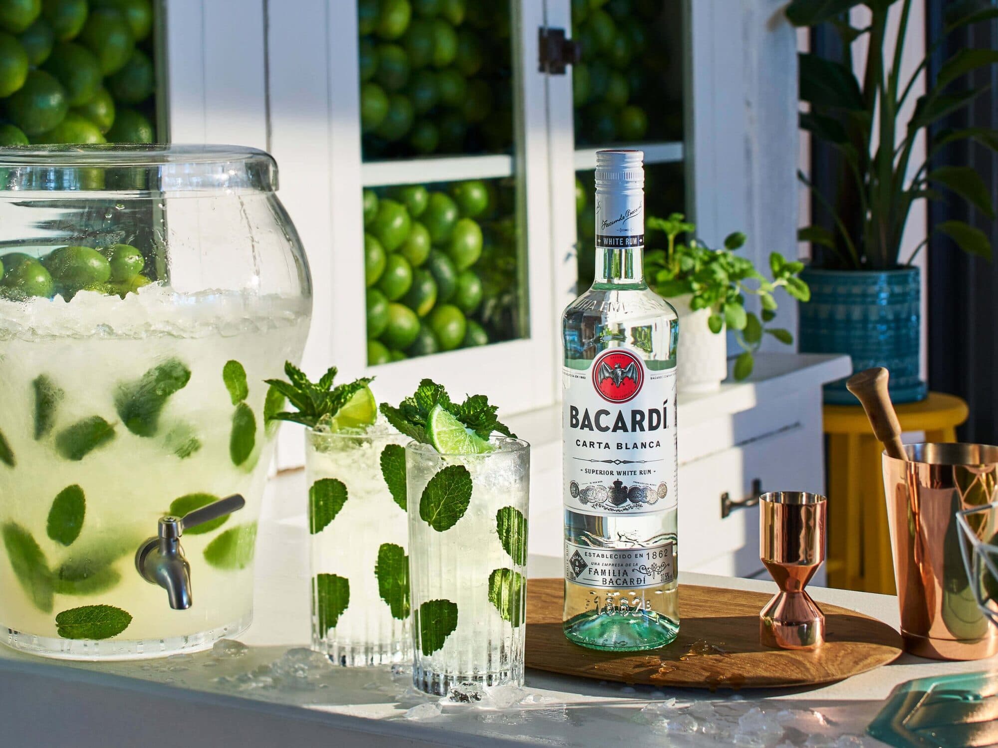 BACARDÍ Carta Blanca rum bottle beside mojito glasses, cocktail tools, large mojito dispenser.