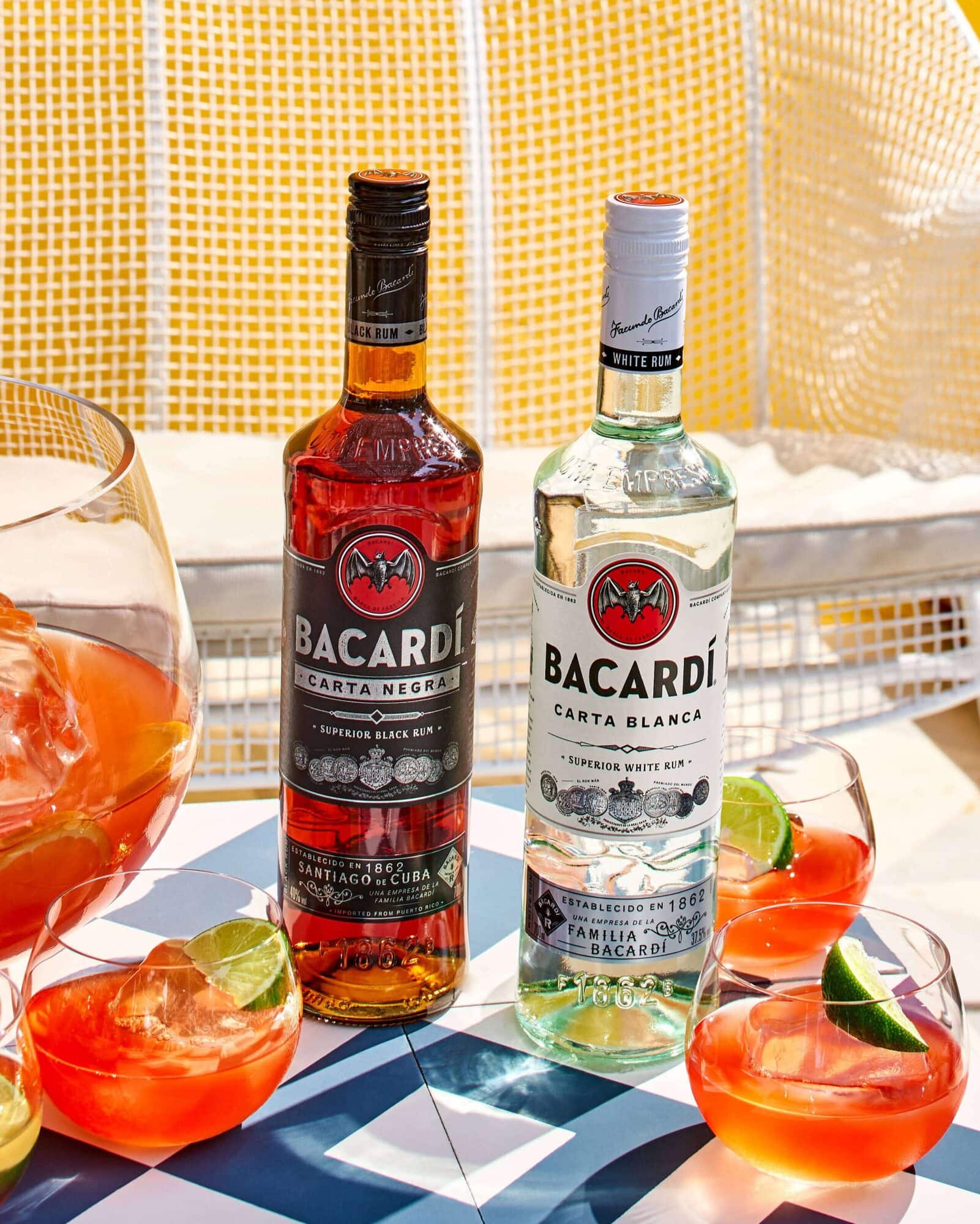 Two BACARDÍ rum bottles, Carta Negra and Carta Blanca, with red-orange cocktails and lime wedges.