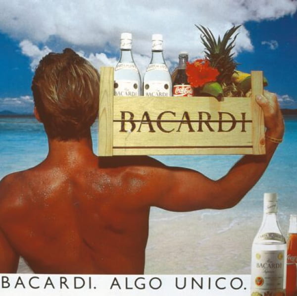 Person carrying wooden crate with rum bottles, pineapple, soda, red flower on a tropical beach.
