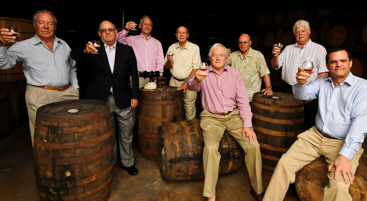 Eight men gathered in a dimly lit cellar, toasting with glasses of amber liquid among wooden barrels.