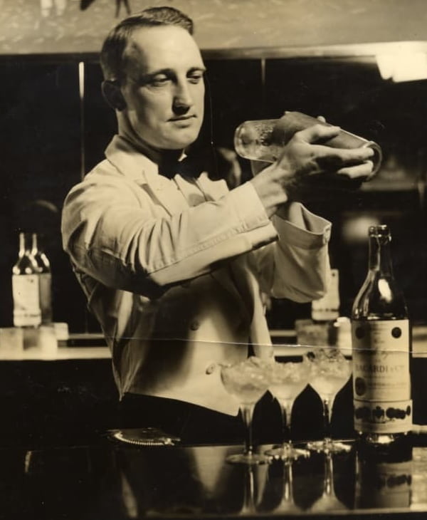 Vintage bartender in white jacket shaking cocktail, three glasses and BACARDÍ bottle on bar top.