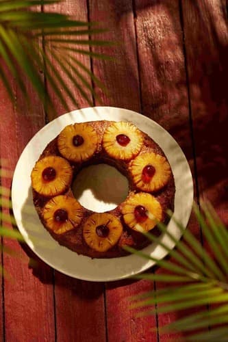 Pineapple upside-down cake with pineapple slices, cherries on white plate under palm shadows.