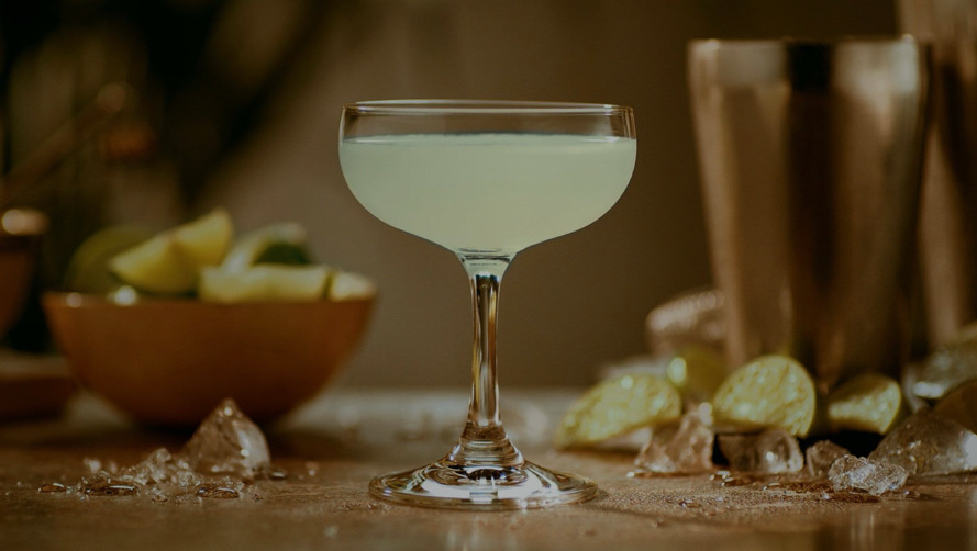 Pale green cocktail in coupe glass, surrounded by lime wedges, ice shards, cocktail shaker.