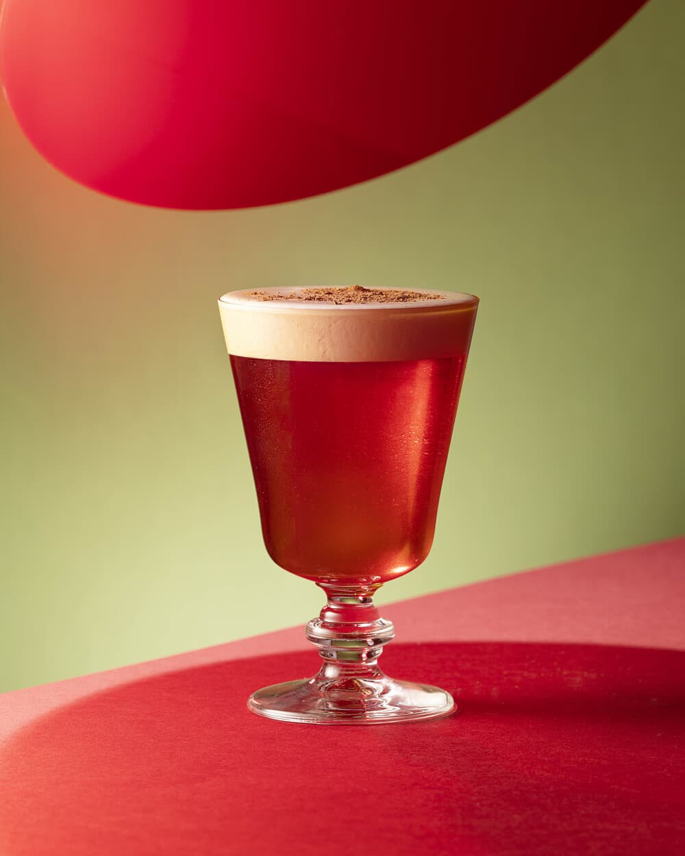 Red cocktail in a stemmed glass with creamy foam head, sprinkled with dark spice.