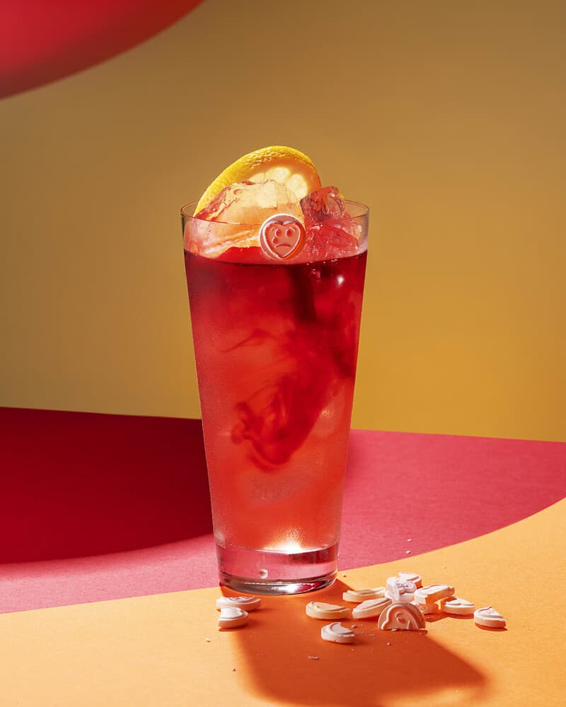 Tall red cocktail with ice, orange slice, heart-shaped candy with sad face.