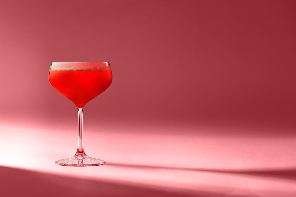Red cocktail in a stemmed coupe glass positioned on a pink background.