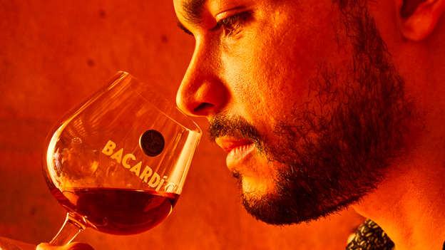 Person in profile smelling a BACARDÍ-branded glass of dark amber liquid.