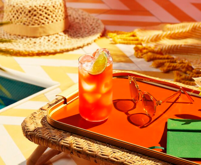 Red cocktail with lime wedge, ice, sunglasses, notebook on an orange tray near a swimming pool.