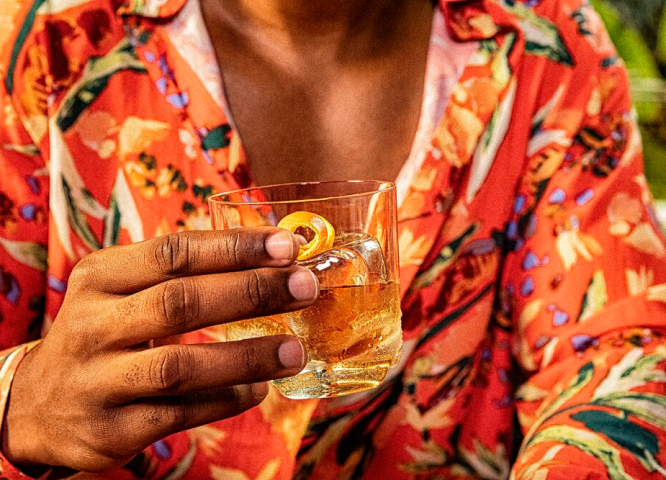 Hand holding clear glass with amber cocktail, large square ice cube, orange peel garnish.