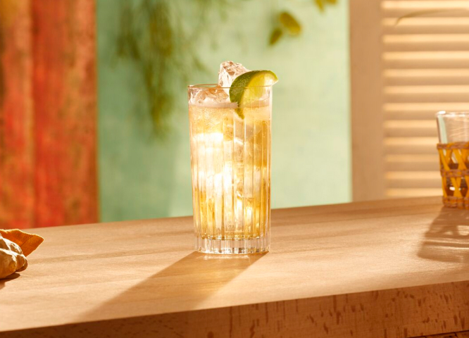 Tall ribbed glass with golden cocktail, ice cubes, fresh lime wedge.