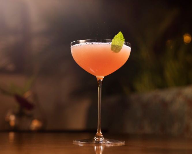 Pinkish-orange cocktail in coupe glass, topped with frothy layer, mint leaf garnish.
