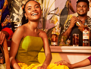 Two people at social gathering, one holding cocktail shaker, BACARDÍ Spiced rum bottles visible.