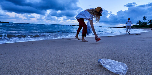 Person bending down to pick up a plastic bottle on a sandy beach during twilight.