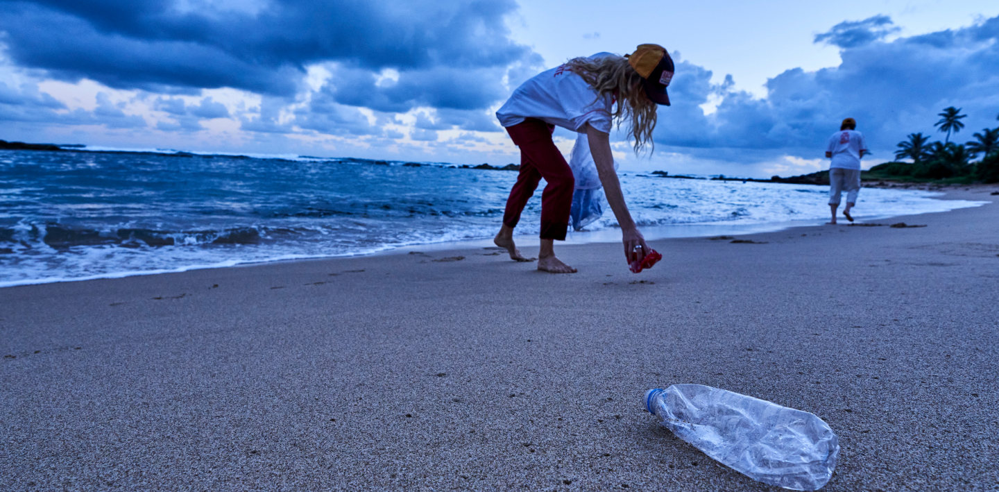 Person bending to collect litter on sandy beach with ocean, clouds, palm trees in background.