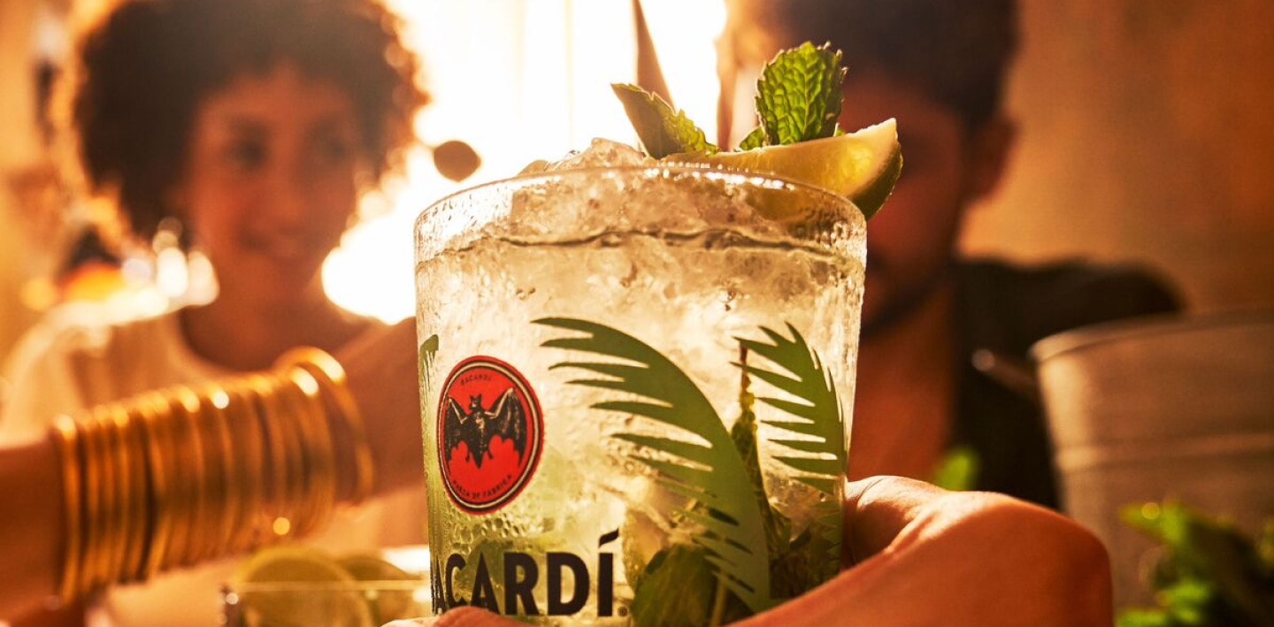 Hand holding a mojito cocktail in a branded glass with mint leaves, lime wedge, crushed ice.
