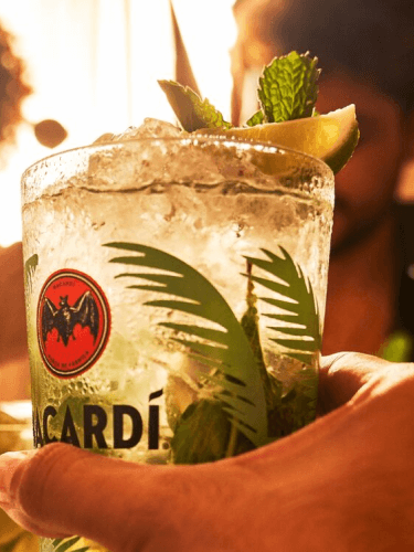 Mojito cocktail in branded glass with crushed ice, mint leaves, lime wedge, held by hand.