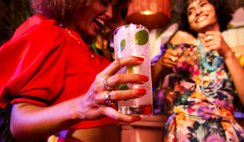 Person holding a tall glass with ice, mint leaves, clear cocktail in a social setting.