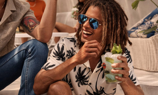 Person holding mojito cocktail, smiling, wearing white shirt with palm tree pattern.