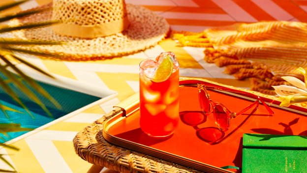 Red cocktail with lime wedge on a tray, accompanied by sunglasses, notebook, poolside accessories.