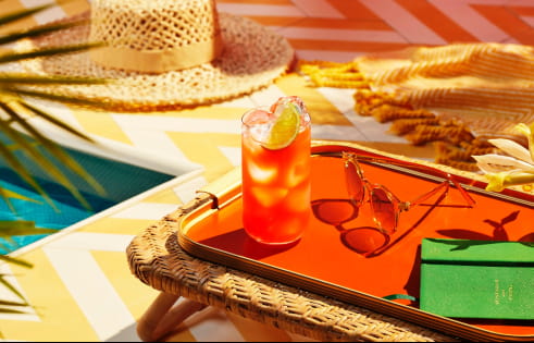 Cocktail with lime wedge on an orange tray, surrounded by sunglasses, notebook, poolside accessories.
