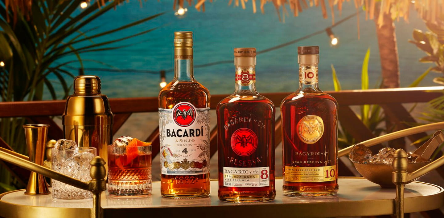 Three BACARDÍ rum bottles arranged on a gold bar cart with cocktail tools and prepared drinks.
