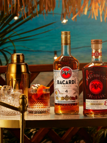 Two BACARDÍ rum bottles, cocktail in rocks glass, gold shaker, empty tumblers arranged on bar surface.