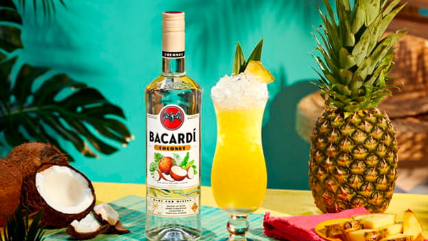 BACARDÍ Coconut rum bottle next to yellow cocktail, with coconut, pineapple, garnished hurricane glass.