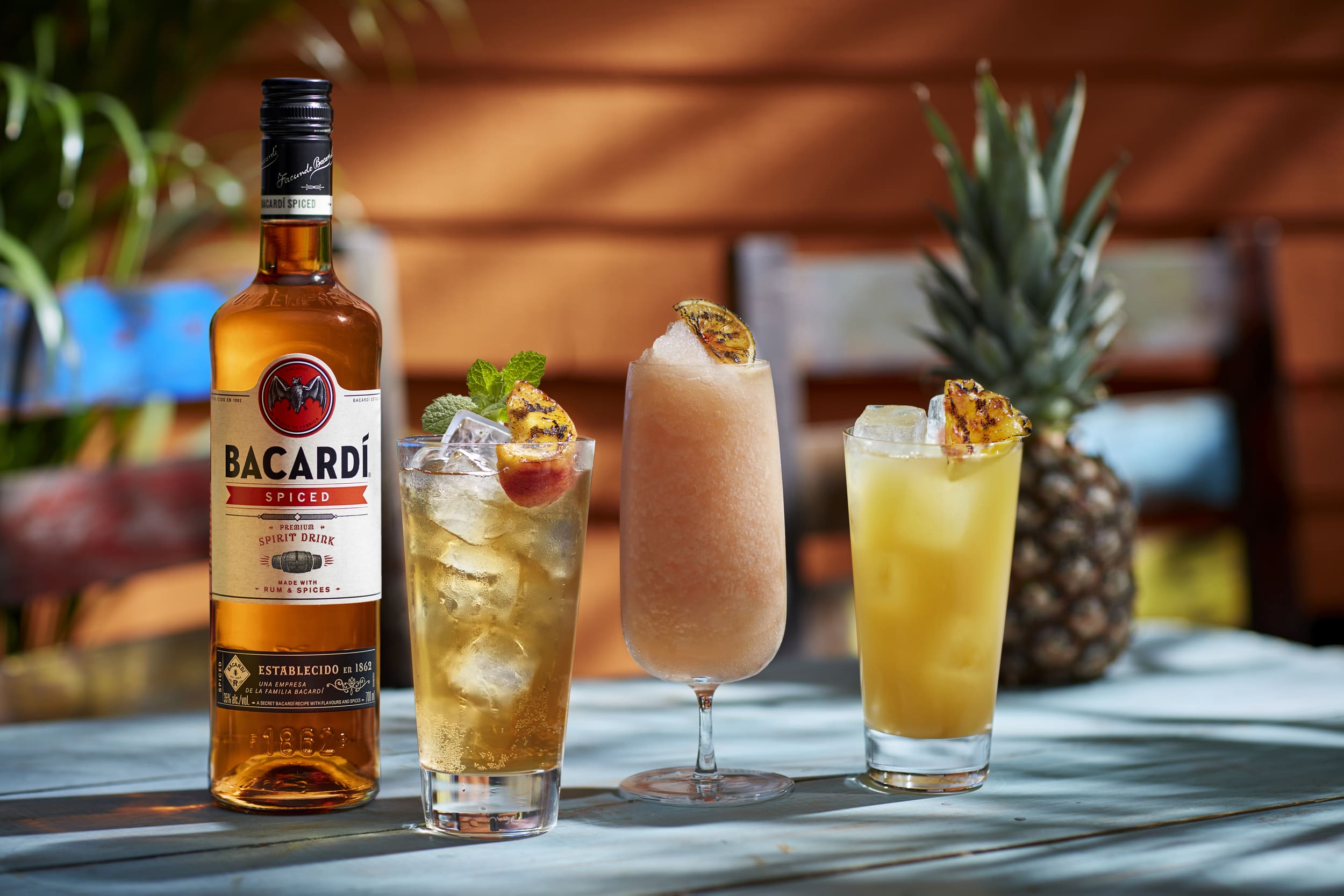 BACARDÍ Spiced rum bottle next to three cocktails garnished with grilled fruit, mint, and whole pineapple.