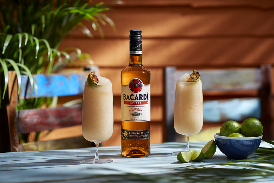 Bacardí Spiced rum bottle between two frosted cocktails with grilled lime wedges and a blue bowl of limes.
