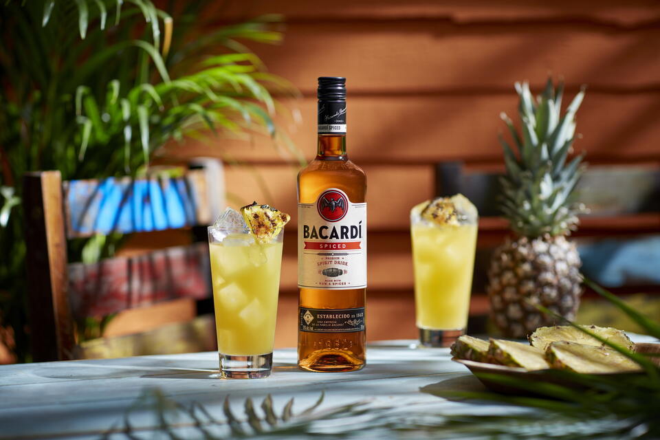 BACARDÍ Spiced bottle with two yellow cocktails, each garnished with grilled pineapple and whole pineapple.