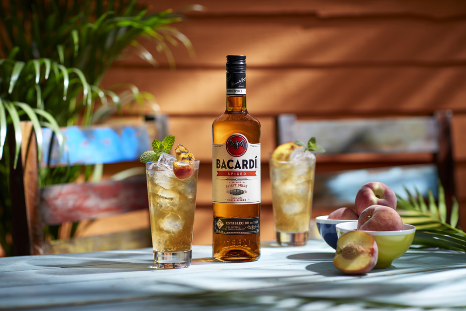 BACARDÍ Spiced rum bottle with two cocktails garnished by grilled peach slices, mint, and fresh peaches in bowls.