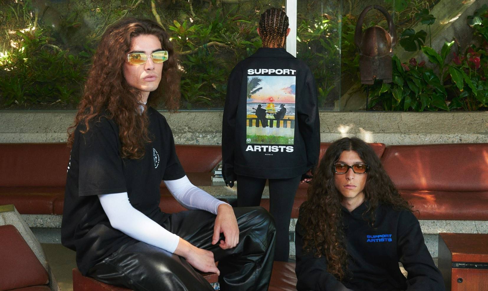 Three people in casual streetwear pose together, showcasing a jacket with a "SUPPORT ARTISTS" graphic print.