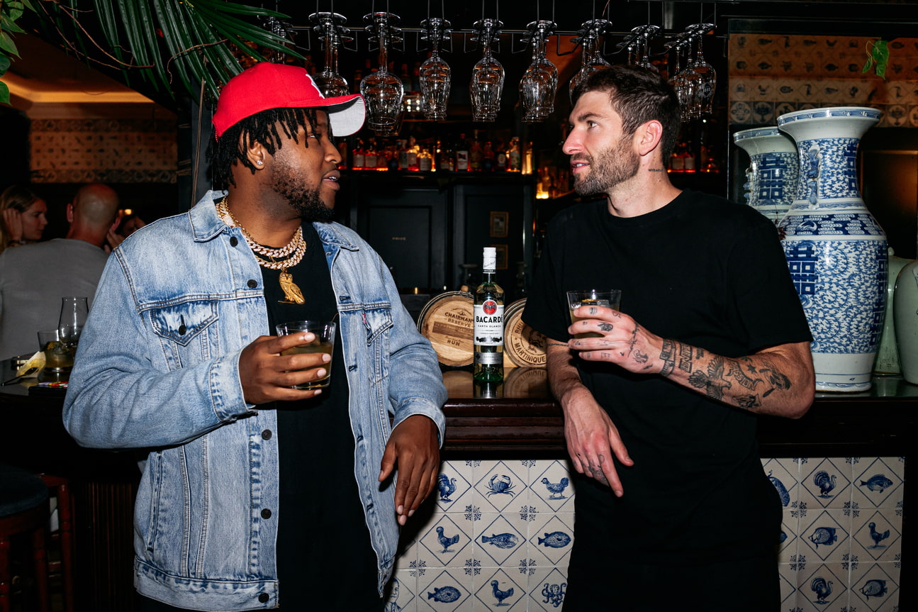 Two men standing at a bar with a Bacardí rum bottle between them, engaged in conversation.