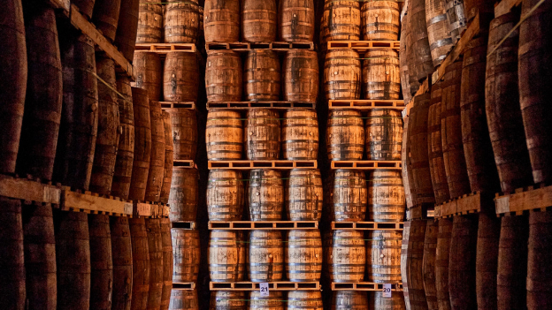 Rows of weathered wooden barrels stacked high on pallets within a dimly lit warehouse space.