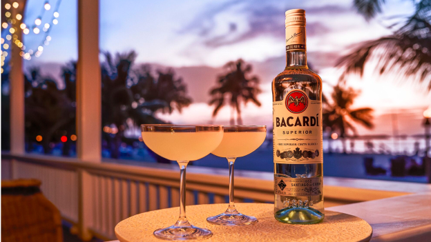 Bacardí Superior rum bottle and two coupe cocktail glasses on a balcony overlooking a sunset beach.