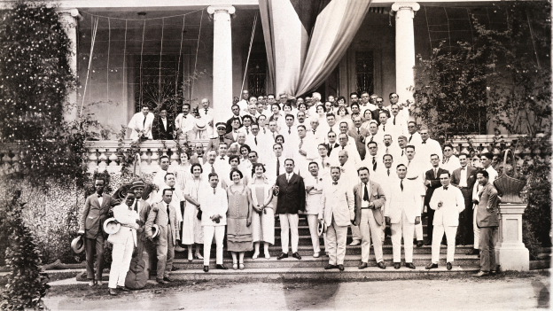 Large group in formal attire posing on building steps with a hanging flag and white columns.