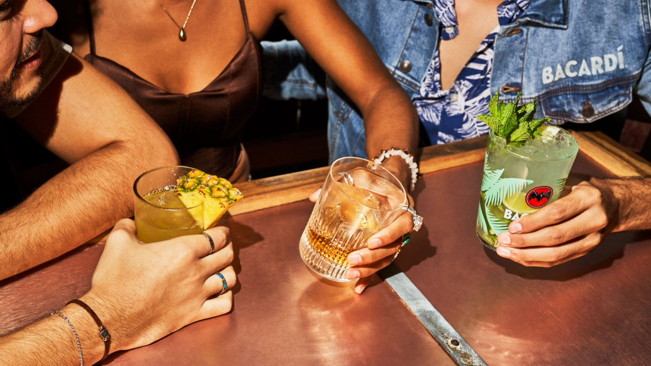 Three hands holding different cocktails: yellow with pineapple, amber in textured glass, green mojito.