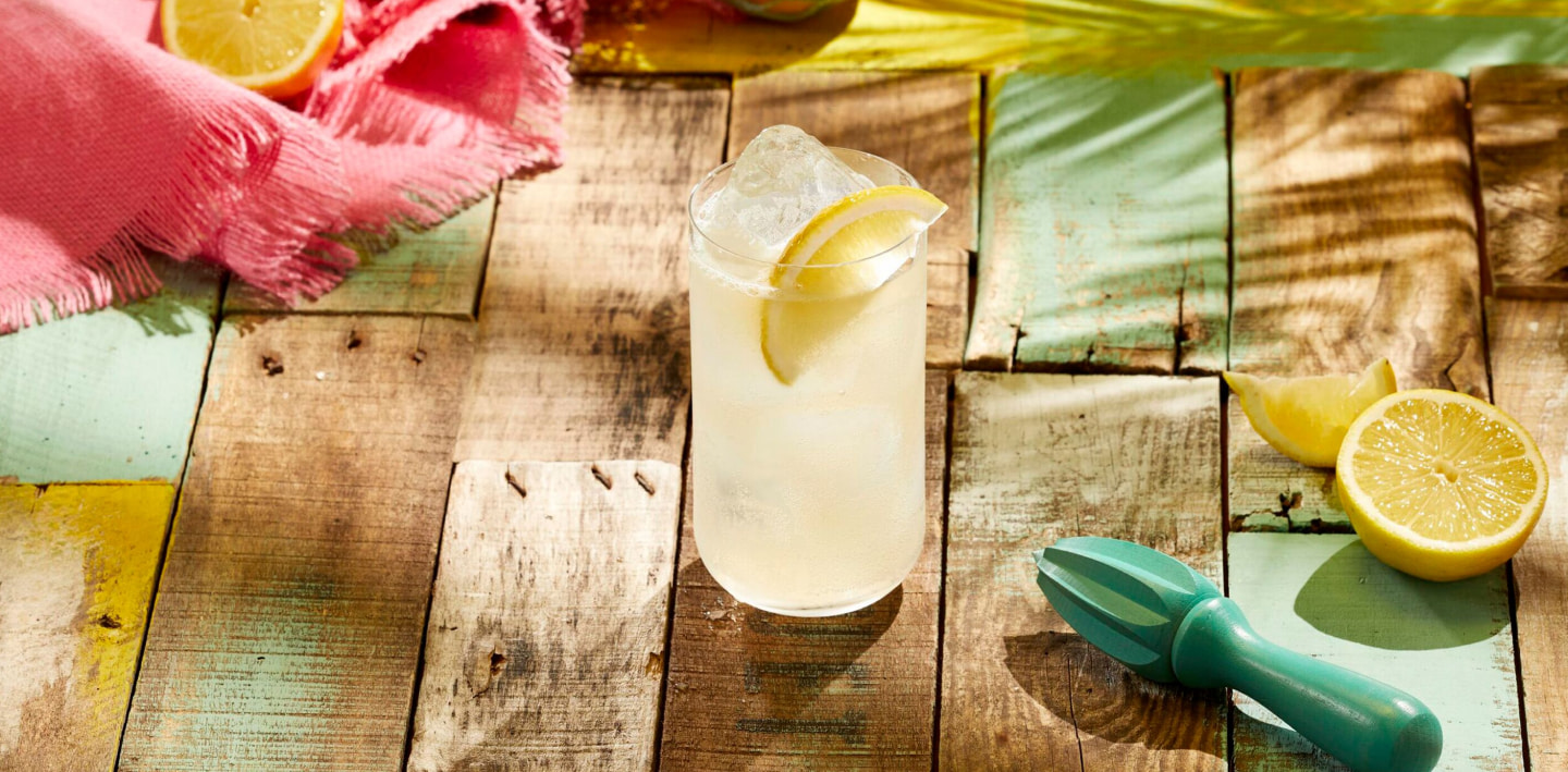 Tall glass of pale yellow cocktail with ice, lemon slice, surrounded by citrus juicer and lemon halves.