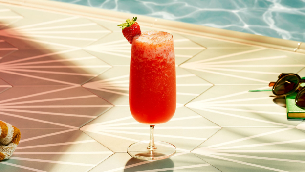 Strawberry slushy cocktail with strawberry garnish, positioned near towel, sunglasses, book by pool.
